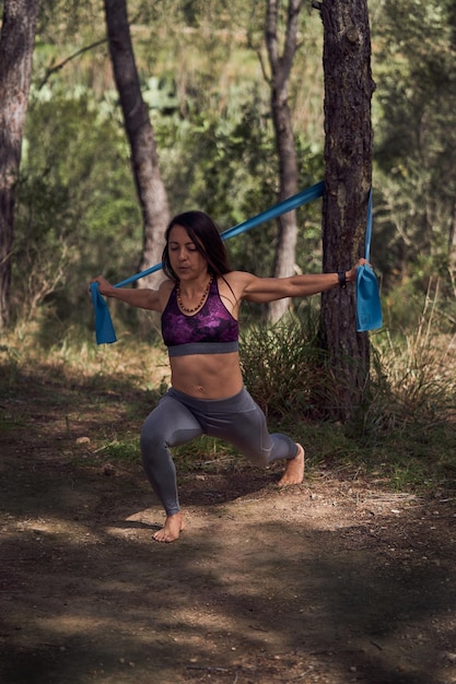 Foto cuerpo completo de mujer hispana descalza vestida con top ajustado y calzas haciendo estocadas con banda de goma de resistencia en el bosque