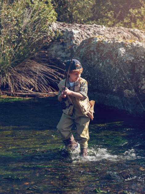 Cuerpo completo de niño concentrado con caña de pescar tratando de sacar peces del agua mientras está parado en el río cerca de la costa