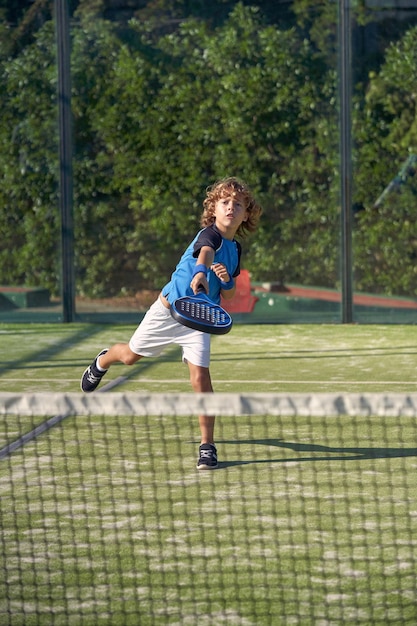Cuerpo completo de niño enérgico en ropa deportiva jugando al pádel con raqueta mientras entrena en el patio de juegos con red en el día de verano