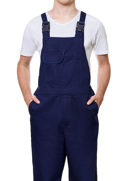 Cuerpo de hombre joven vestido con un mono azul oscuro y una camiseta blanca con las manos en los bolsillos, aislado en la pared blanca.