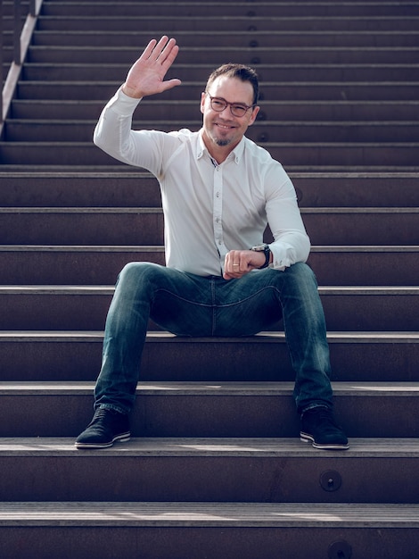 Cuerpo lleno de macho barbudo positivo con gafas agitando la mano y mirando hacia otro lado mientras está sentado en una escalera de piedra en la ciudad