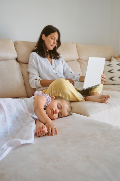 Cuerpo lleno de una mujer joven navegando por la computadora portátil mientras está sentada en un sofá acogedor cerca de la hija durmiente en la sala de estar en casa
