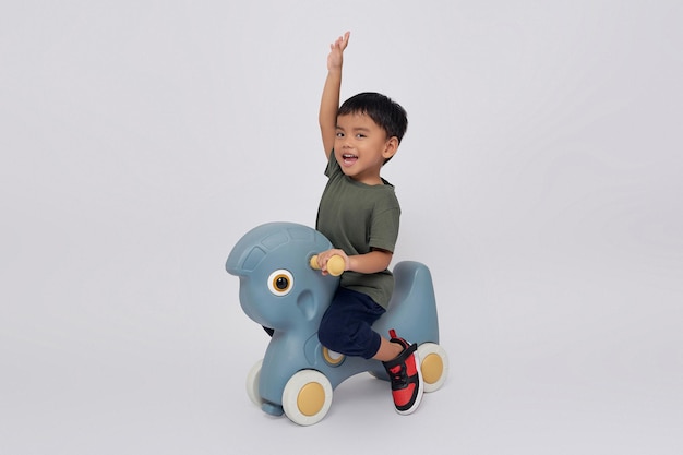 Foto cuerpo lleno pequeño pequeño sonriente feliz chico asiático de 23 años con camiseta verde sentado en un caballo de balanceo de juguete y mostrando el gesto ganador puño cerrado aislado sobre fondo blanco