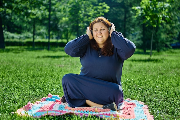 El cuerpo de la mujer de talla grande se sienta en la estera en medio del césped y escucha música con auriculares inalámbricos