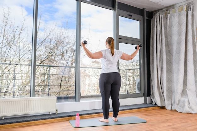 Cuerpo positivo mujer gordita quedarse atrás en leggings negros y levantar las manos con pesas pesas descalzo sobre la alfombra