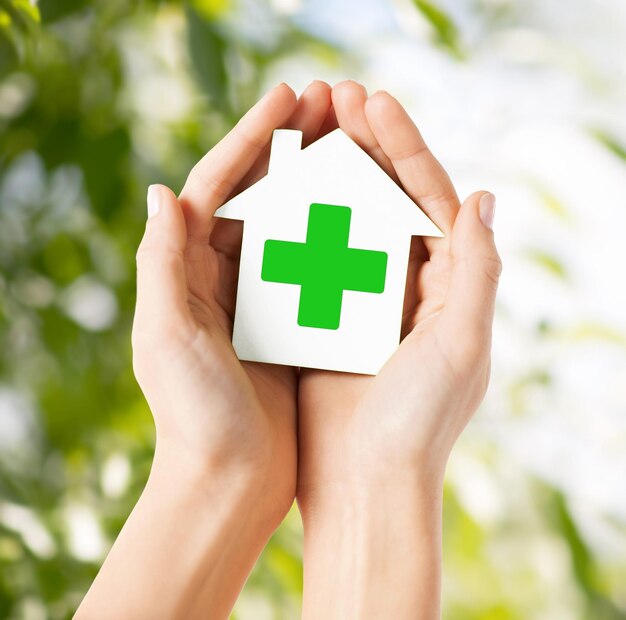 cuidado, ajuda, caridade e conceito de pessoas - close-up de mãos segurando uma casa de papel branco com sinal de cruz verde sobre fundo natural