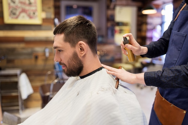 cuidado del cabello, peluquería y concepto de personas - peluquero aplicando spray de peinado a un cliente masculino en la barbería