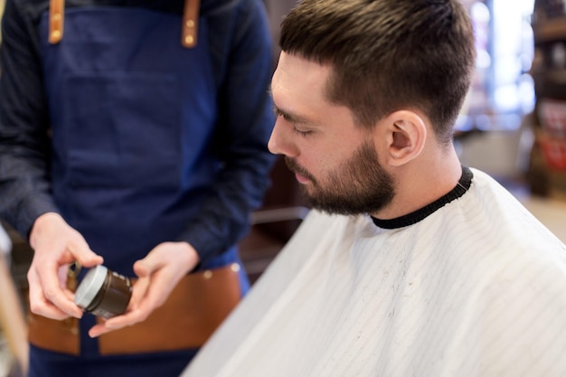 Foto cuidado del cabello, peluquería y concepto de personas - peluquero mostrando cera de peinado a un cliente masculino en la barbería