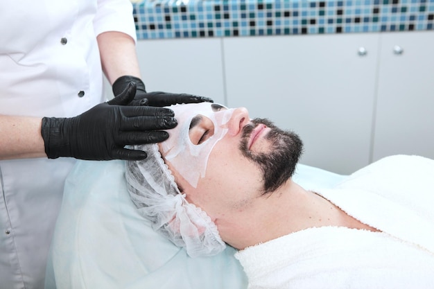 Foto cuidado de la cara del hombre en el salón de belleza hombre con máscara de arcilla en la cara cosmetología de los hombres cosmetóloga pone máscara en la cara en el spa