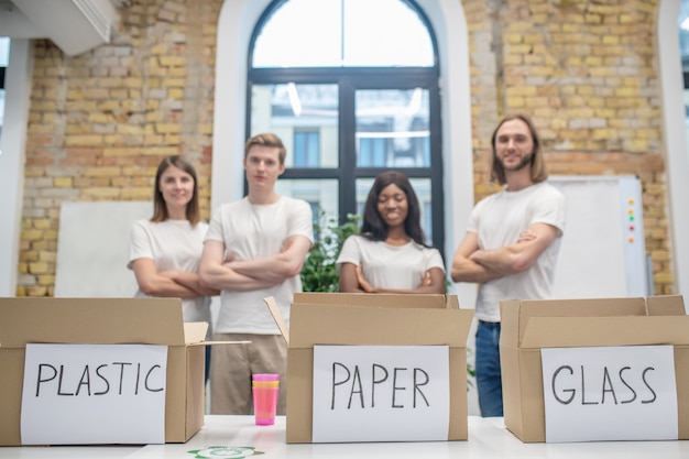 Cuidado, ecologia. Caixas de lixo de papelão com letras na mesa e jovens pessoas satisfeitas confiantes em camisetas brancas atrás