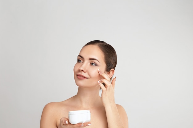 Cuidado facial. Mujer aplicando crema y sonriendo. Retrato de mujer joven con crema cosmética en la piel.