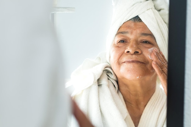 Cuidado facial Mujer mayor sonriendo aplicando crema facial frente al espejo