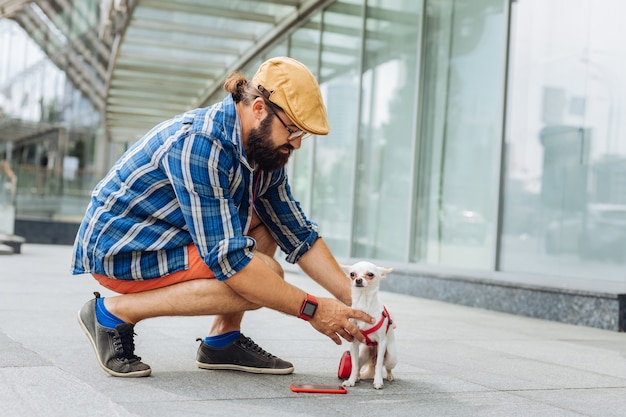 Cuidado del perro. Hombre moreno barbudo vestido con reloj inteligente rojo cuidando de perrito blanco