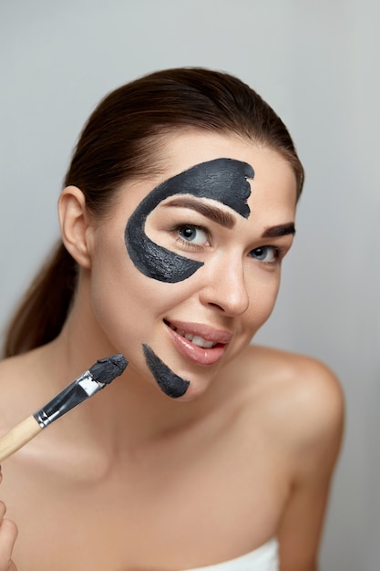 Cuidado de la piel facial de belleza. Mujer sonriente aplicar mascarilla facial de arcilla para el cuidado de la piel. Modelo de niña con máscara cosmética. Tratamiento facial