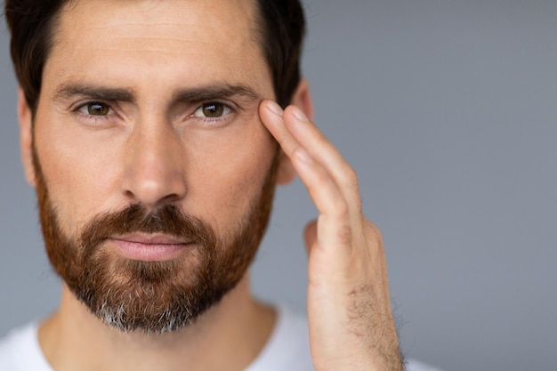 Cuidado de la piel facial masculino Hombre barbudo de mediana edad tocando la cara cuidando la piel debajo de los ojos espacio de copia de fondo gris