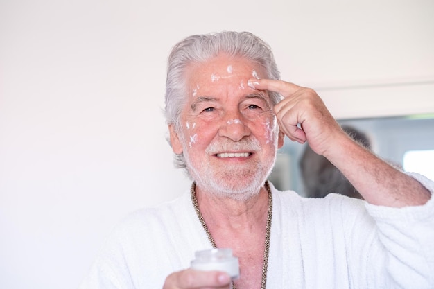 Cuidado de la piel Guapo anciano barbudo en bata de baño aplicando crema en la cara como rutina diaria sosteniendo un frasco de crema