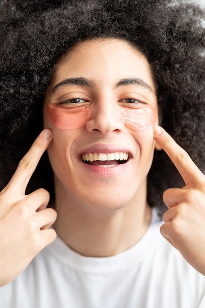 Cuidado de la piel del hombre Producto cosmético Aseo masculino divertido Chico guapo feliz con una sonrisa con dientes cabello rizado marrón apuntando a parches oculares de hidrogel rosa en la piel perfecta de la cara