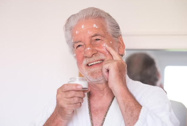 Cuidado de la piel para hombres Hombre guapo con barba senior en bata de baño aplicando crema en la cara como rutina diaria sosteniendo un frasco de crema