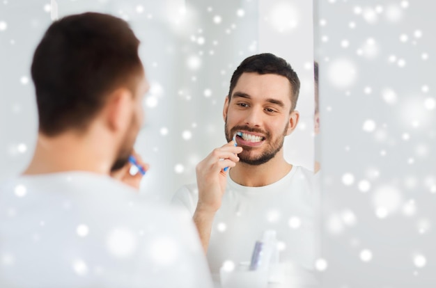 cuidado de la salud, higiene dental, gente y concepto de belleza - joven sonriente con cepillo de dientes limpiando dientes y mirando al espejo en el baño de casa sobre la nieve