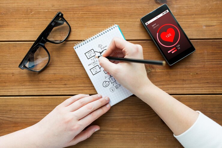 Foto cuidado de la salud, planificación y concepto de personas: cierre del esquema de dibujo de mujeres o estudiantes en el bloc de notas con lápiz y anteojos con frecuencia cardíaca en el teléfono inteligente en la mesa de madera