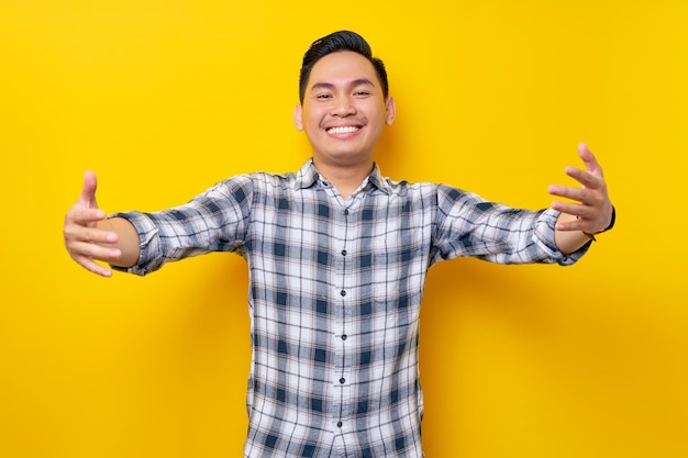 Cuidado sonriente joven apuesto hombre asiático con una camisa a cuadros De pie con las manos extendidas para abrazos aislado sobre fondo amarillo Concepto de estilo de vida de la gente