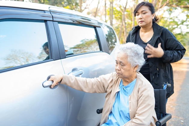El cuidador ayuda y apoya a una paciente mayor asiática sentada en silla de ruedas a prepararse para llegar a su automóvil.