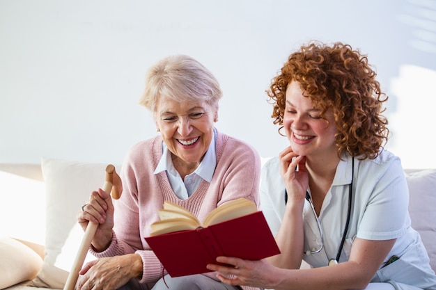 Cuidador de mulher que lê um livro ao sentar-se com a mulher sênior feliz no lar de idosos.