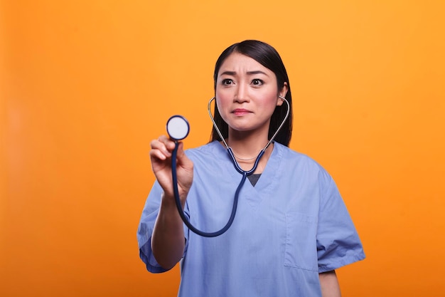 Cuidador de saúde asiático preocupado usando estetoscópio para consultar o paciente em fundo laranja. Enfermeira preocupada com instrumento médico profissional que consulta a pessoa doente.
