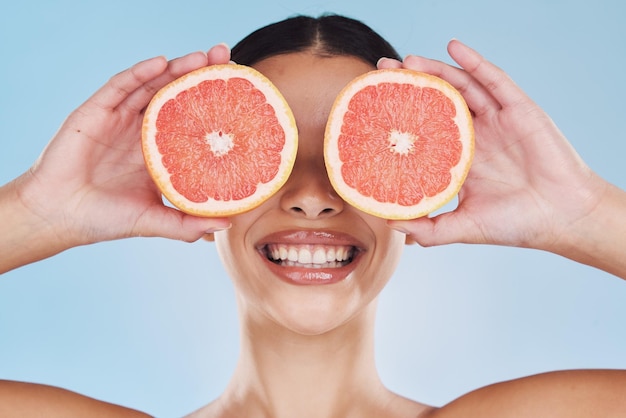 Cuidados com a pele da mulher toranja e pele do rosto com frutas para a saúde do bem-estar e facial em um fundo azul no estúdio Sorriso feliz e modelo de beleza com produto alimentar nas mãos, escondendo os olhos com vitamina c