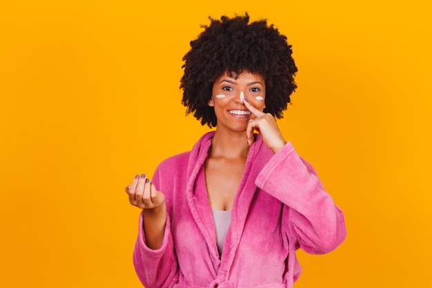 Cuidados com a pele. Mulher afro aplicando creme cosmético no rosto isolado em fundo amarelo