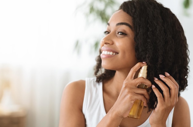 Cuidados com o cabelo mulher afro feliz usando óleo para pontas divididas enquanto se prepara em casa imagem em close-up com