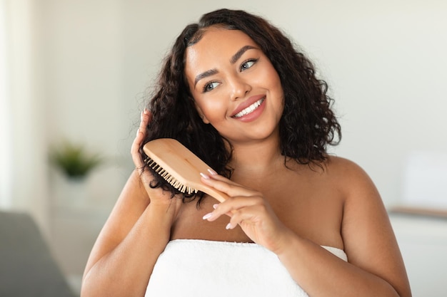 Foto cuidados com os cabelos domésticos feliz mulher afro-americana corporal positiva escovando o cabelo com escova em pé no interior do banheiro