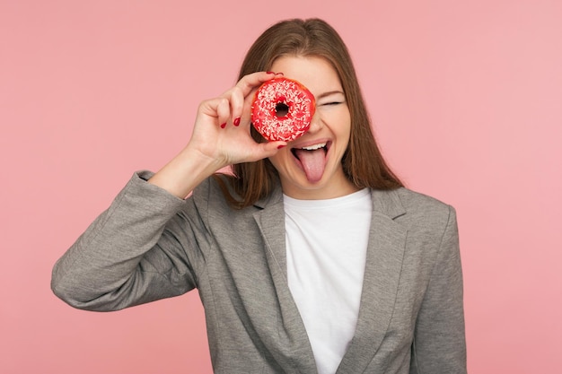 Cuidados de saúde e nutrição. Retrato de empresária alegre de paletó olhando através de rosquinha doce, se divertindo com pastelaria, espiando rosquinha e sorrindo. tiro de estúdio interno, fundo rosa