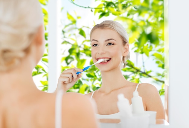 cuidados de saúde, higiene dental, pessoas e conceito de beleza - jovem sorridente com escova de dentes limpando os dentes e olhando para o espelho no banheiro de casa sobre fundo verde natural