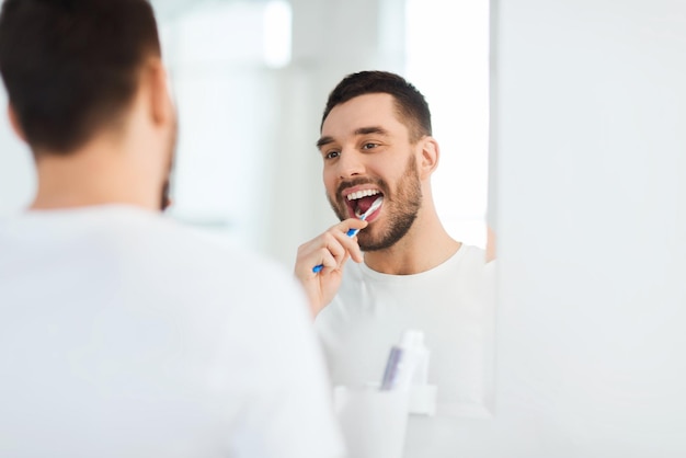 cuidados de saúde, higiene dental, pessoas e conceito de beleza - jovem sorridente com escova de dentes limpando os dentes e olhando para o espelho no banheiro de casa