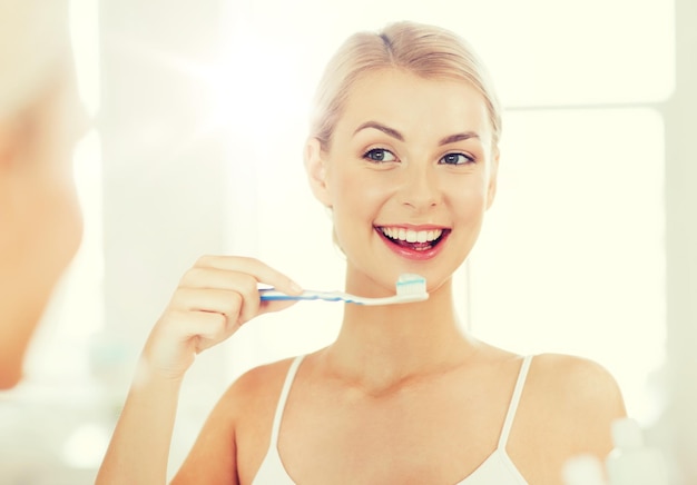 cuidados de saúde, higiene dental, pessoas e conceito de beleza - jovem sorridente com escova de dentes limpando os dentes e olhando para o espelho no banheiro de casa