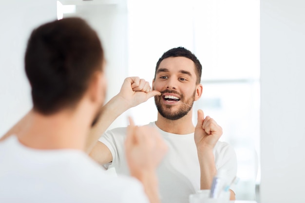 cuidados de saúde, higiene dental, pessoas e conceito de beleza - jovem sorridente com fio dental limpando os dentes e olhando para o espelho no banheiro de casa