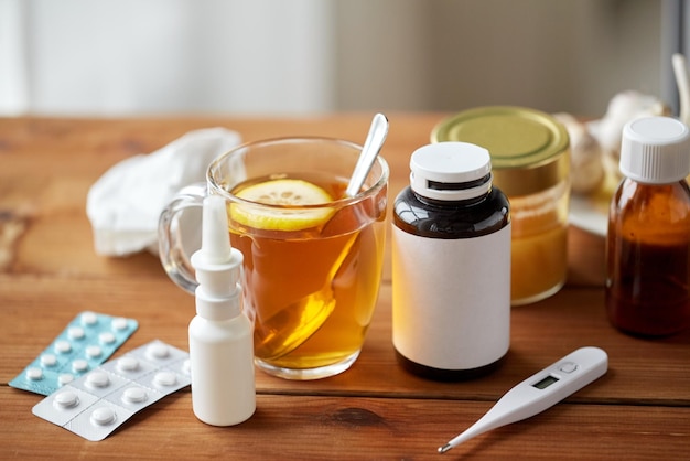 cuidados de saúde, medicina tradicional e conceito de gripe - xícara de chá com limão, termômetro e drogas na mesa de madeira