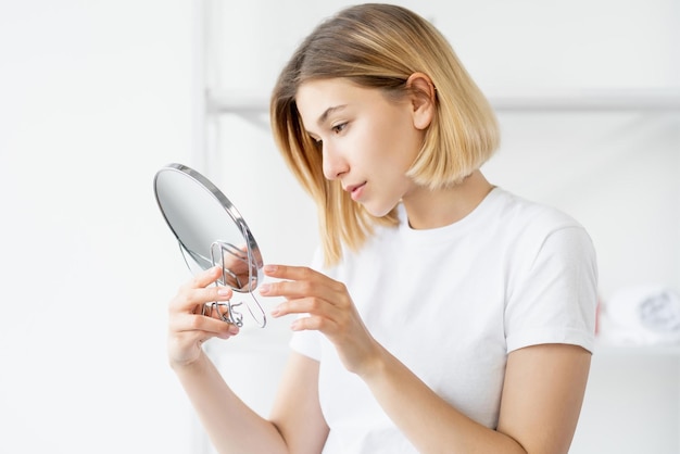 Cuidados matinais no banheiro Tratamento facial Beleza bem-estar Mulher atraente fresca admirando a pele perfeita do rosto no espelho na sala interna de luz desfocada