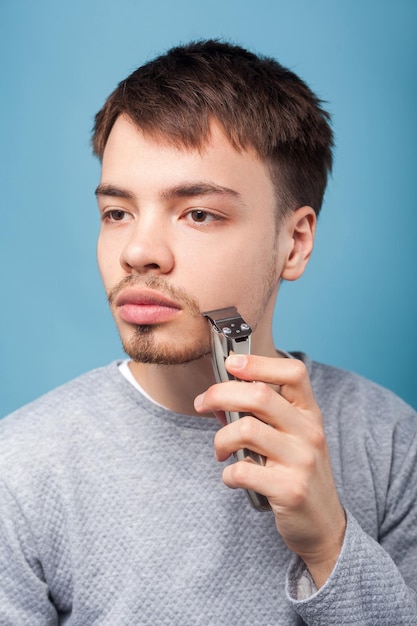 Cuidados pessoais e cuidados pessoais Retrato de homem moreno concentrado concentrado fazendo a barba com aparador ou barbeador elétrico olhando de lado com sério rosto atento studio tiro isolado no fundo azul