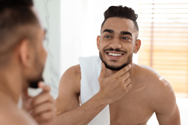 Cuidados pós-barba homem árabe sem camisa bonito com toalha no ombro olhando no espelho