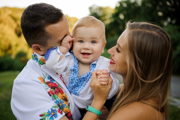 Cuidadosos padres sosteniendo en las manos a un bebé sonriente vestido con la camisa bordada