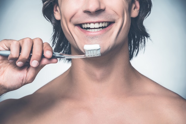 Cuidando bien mis dientes. Primer plano de apuesto joven sin camisa cepillándose los dientes y sonriendo mientras está de pie contra el fondo gris