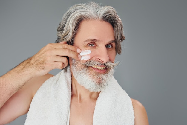 Cuidando la claridad de la cara y la piel El hombre mayor moderno y elegante con cabello gris y barba está en el interior