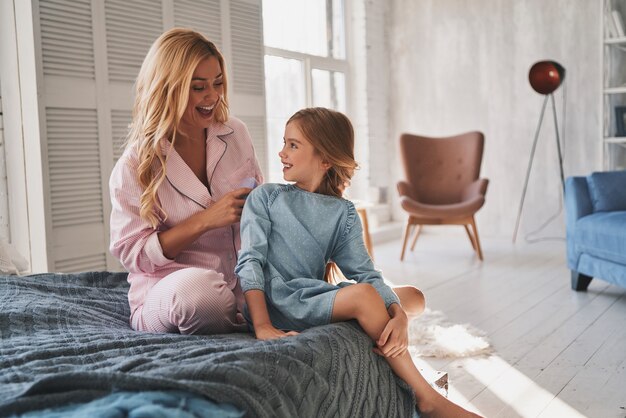 Cuidándola bien. Hermosa joven madre cepillando el cabello de su hija mientras está sentada en la cama en casa