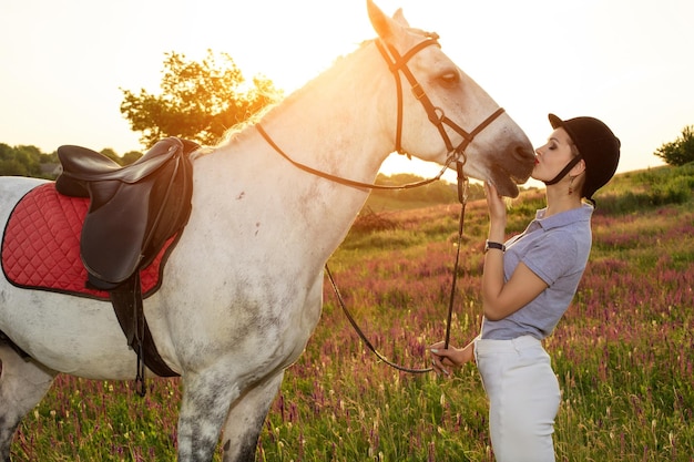 Cuidar de los animales, el concepto de amor y amistad. Joven jockey acariciando y abrazando a un caballo blanco al atardecer