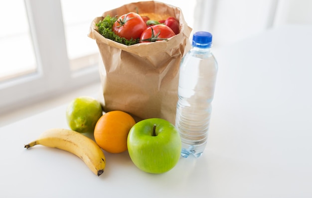 culinária, dieta, comida vegetariana e conceito de alimentação saudável - close-up do saco de papel com frutas suculentas frescas e legumes e garrafa de água na mesa da cozinha em casa