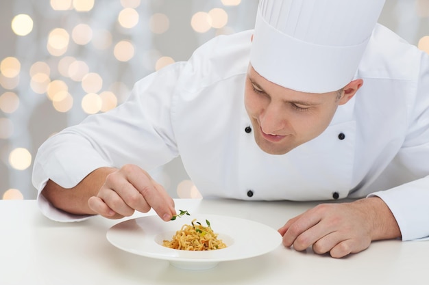 culinária, profissão, alta cozinha, comida e conceito de pessoas - feliz chef masculino cozinhar prato de decoração sobre fundo de luzes de férias de natal