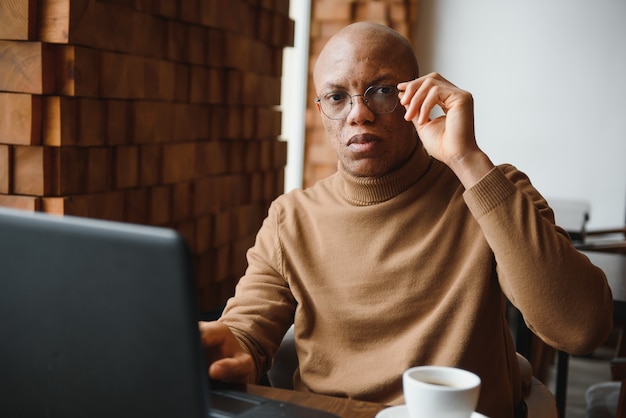 Óculos de estudante do sexo masculino negro sério tomando notas enquanto está sentado à mesa com o laptop e trabalhando em um projeto em um café aconchegante.