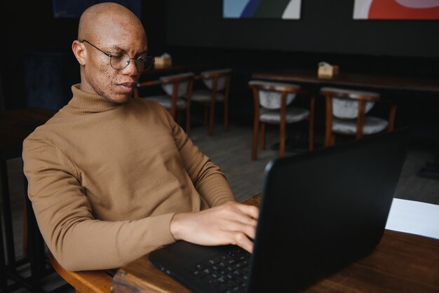 Óculos de estudante do sexo masculino negro sério tomando notas enquanto está sentado à mesa com o laptop e trabalhando em um projeto em um café aconchegante.